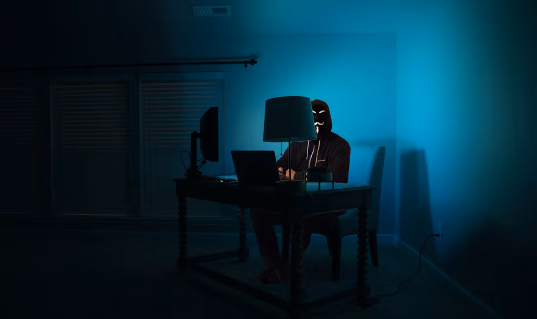 A hooded figure sits in front of a laptop screen in a darkened room. (Kartik Programmer/Unsplash)